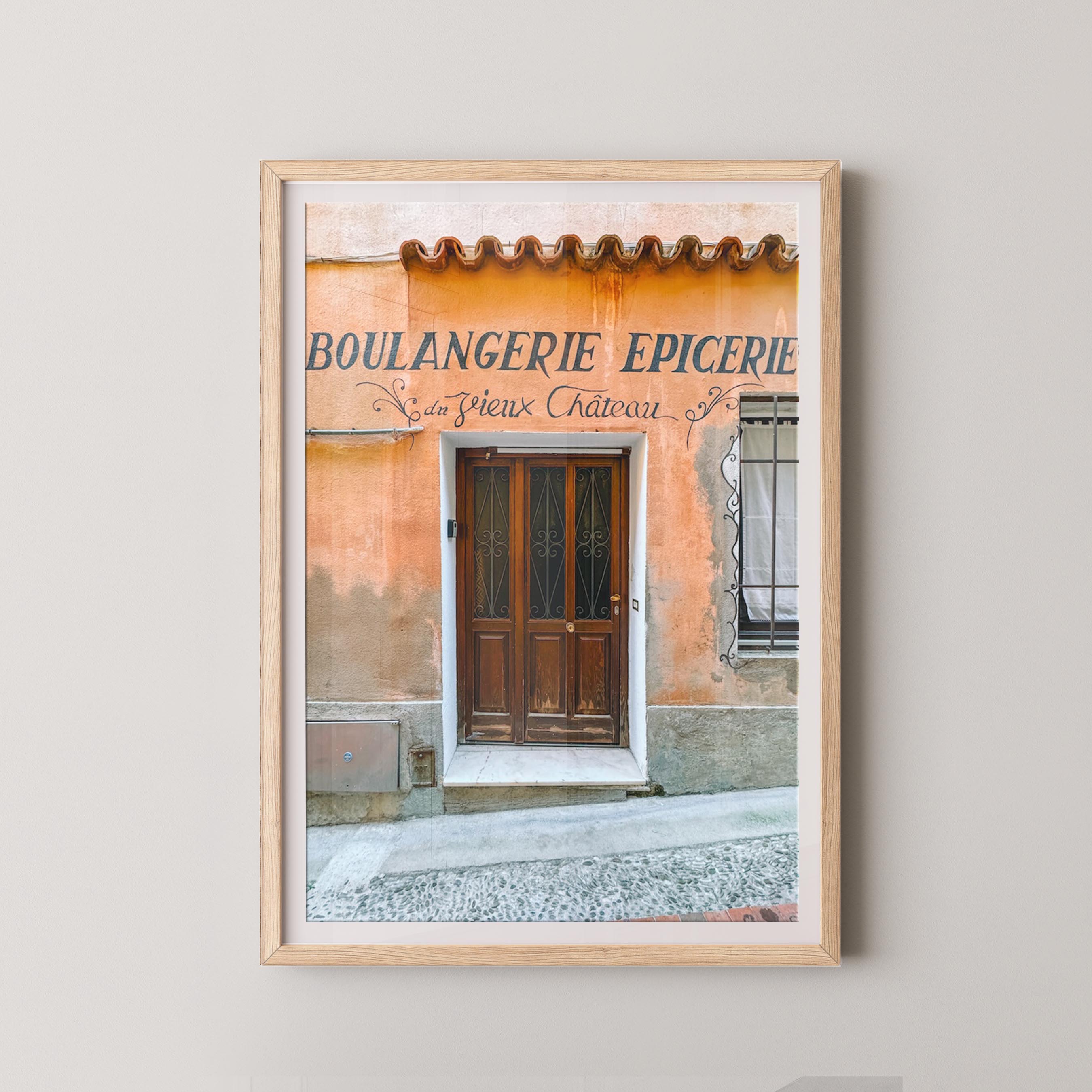 Framed print of a bakery storefront with a wooden door and sign on a light gray wall.